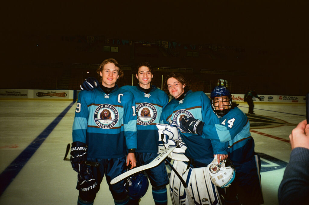 "Four hockey players in blue uniforms posing together on an ice rink, smiling for a photo. The rink is indoors with boards and advertisements visible in the background, suggesting a team game or practice session."