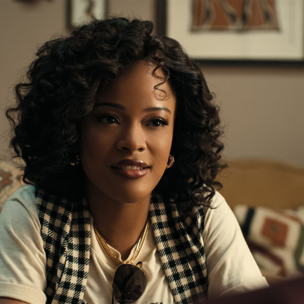 "A young woman with curly hair sits on a patterned couch in a cozy living room, wearing a checkered vest and white shirt, with framed art and cushions in the background, engaged in conversation."